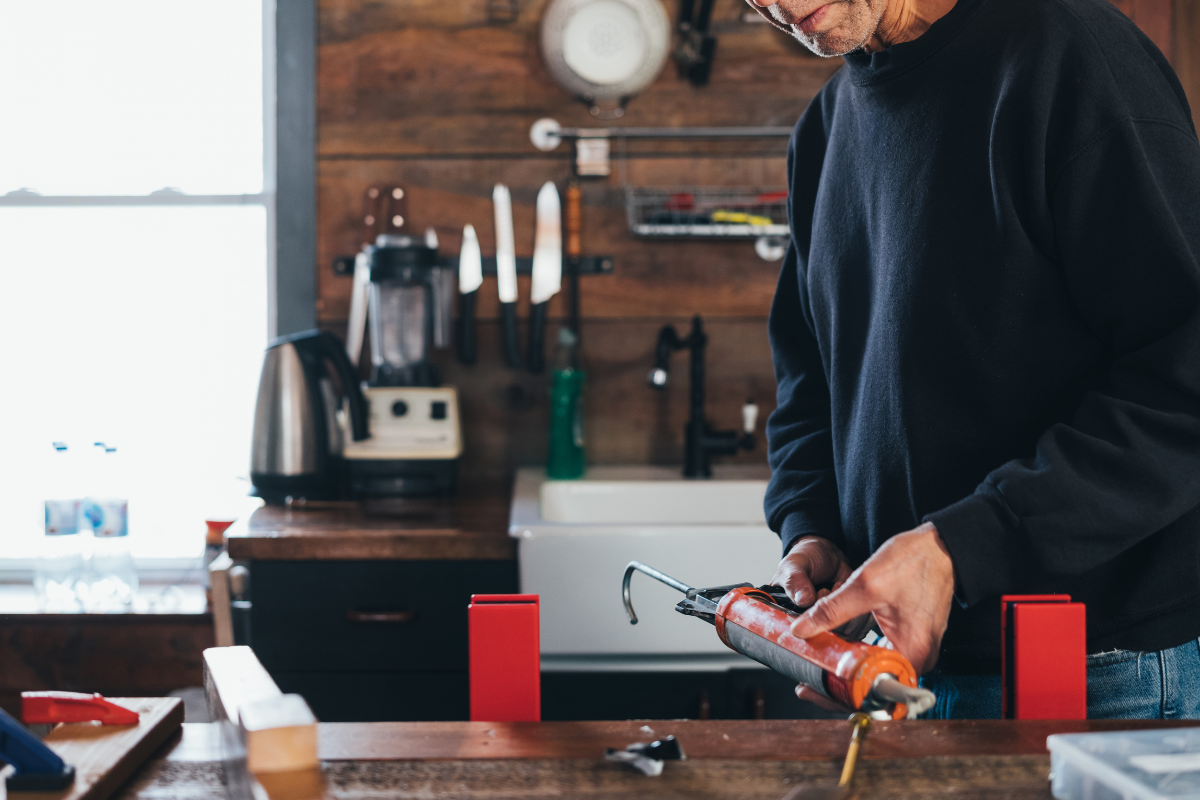 Person in a workshop holding a caulking gun near a workbench with tools mounted on a pegboard